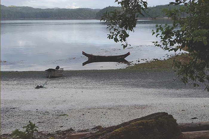 View of a cove when the tide is low. There is an aluminum boat tied to a log and resting on the shore. There's also a carved canoe sitting in the water.