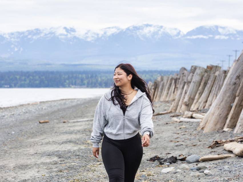 A student walking along Goose Spit beach in Comox, on a sunny day. There are mountains and water in the background.