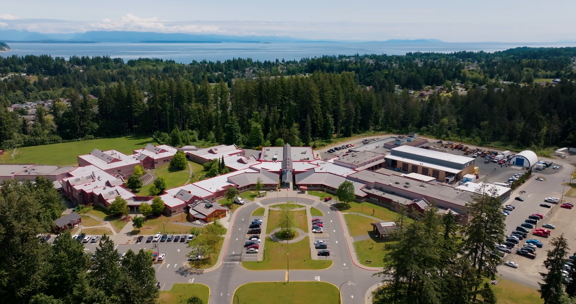 Aerial image of Campbell River campus on a sunny day, with trees, mountains and ocean in the distance.
